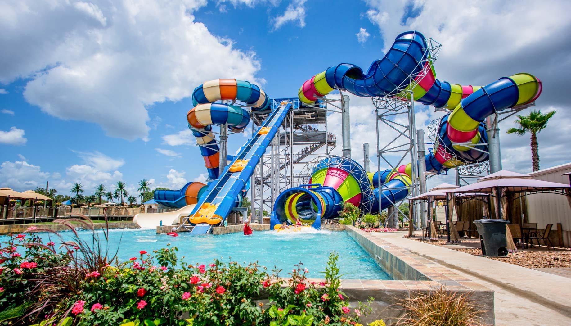 A colorful water park features a large, winding waterslide ending in a clear blue pool surrounded by deck chairs, cabanas, and palm trees under a partly cloudy sky.