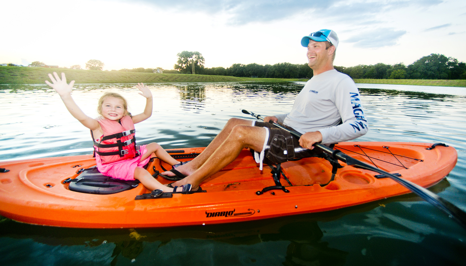 An adult and a child, both smiling and waving, are seated in an orange kayak on a calm body of water surrounded by trees.