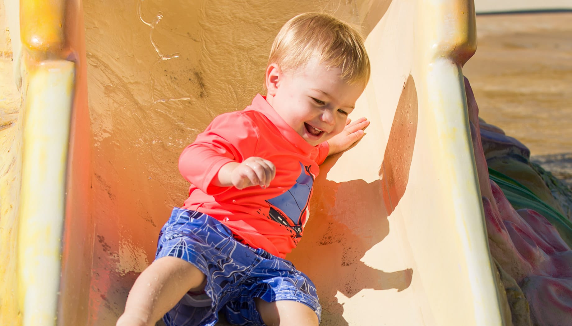 Toddler boy in a red shirt and blue swim trucks sliding down a small water slide.