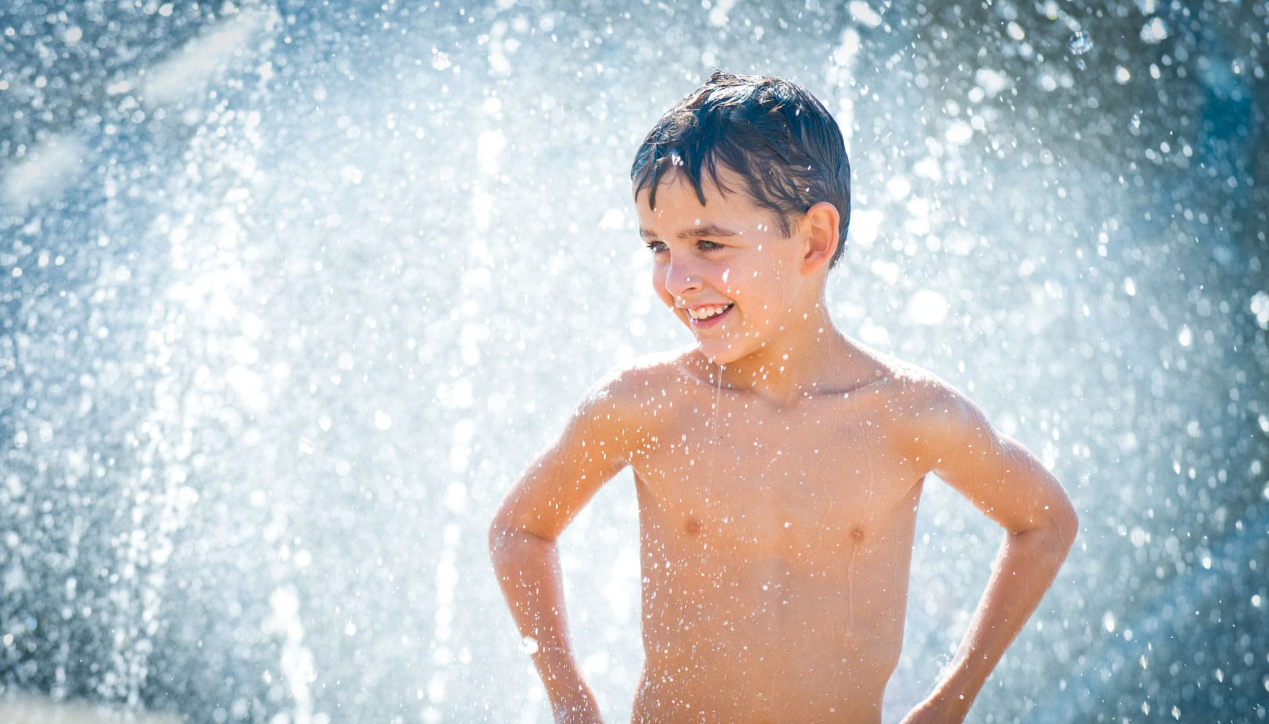 A boy stands with hands on hips, smiling under a spray of water, with droplets glistening around him.