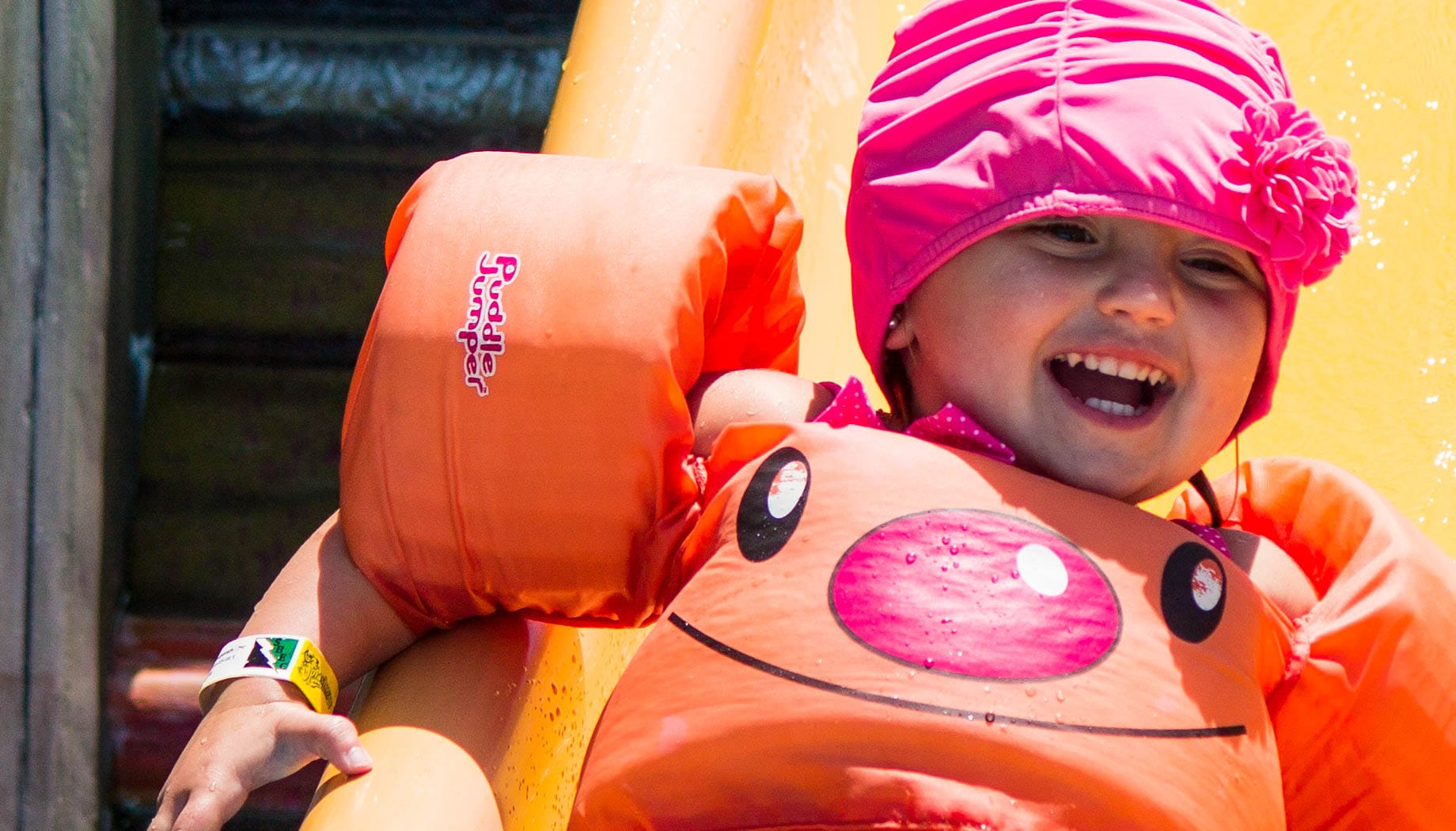 A young child in orange floaties and a pink swim cap smiles while sliding down a water slide.