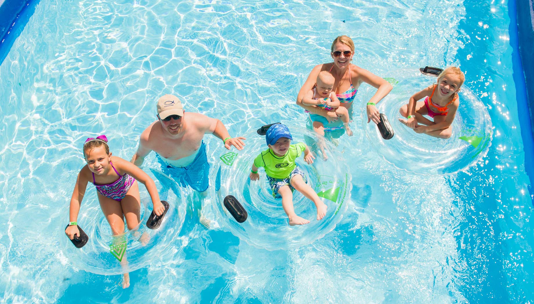 A group of five people, two adults and three children, float in a lazy river using inflatable rings on a sunny day.