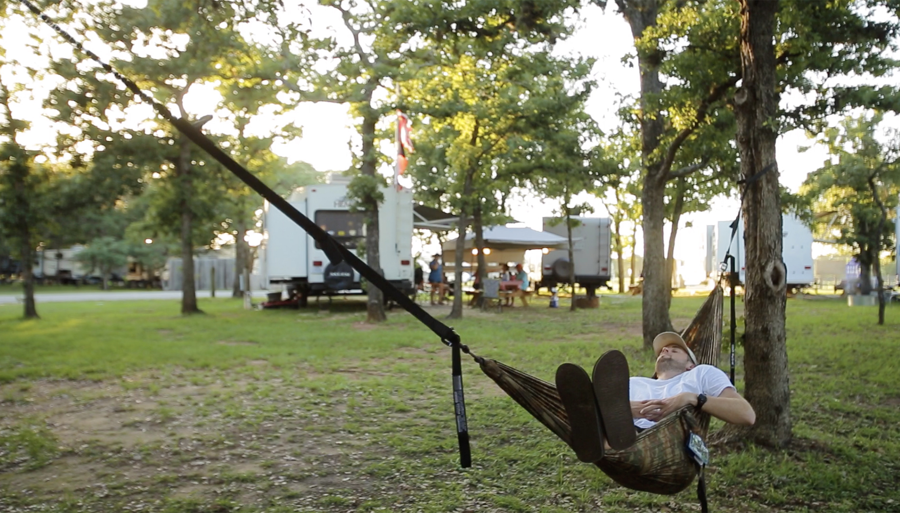 A person relaxes in a hammock tied between two trees in a campsite with parked RVs and people in the background.