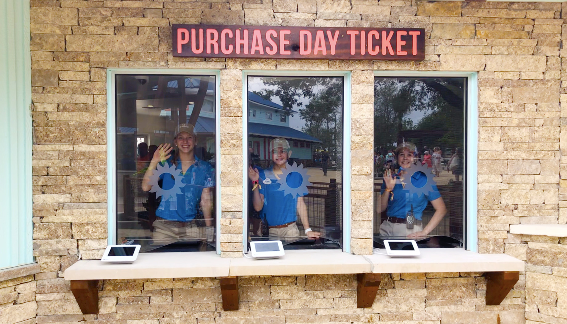 Three ticket booth attendants in blue shirts wave from behind glass windows at a ticket counter with a "Purchase Day Ticket" sign above.