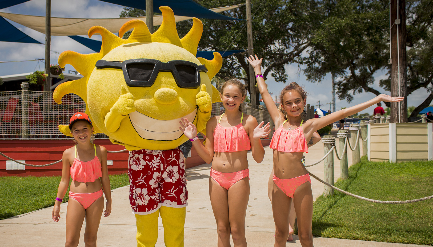 Three children in pink swimsuits pose with a person in a large sun mascot costume dressed in a red Hawaiian shirt with sunglasses, under sunny outdoor conditions.