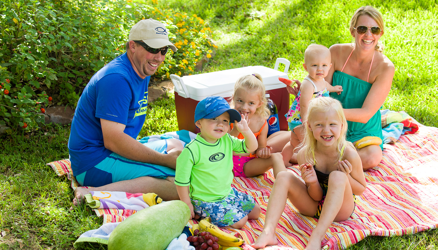 A family enjoys a picnic on a colorful blanket. Two adults and four children are sitting on the grass with a cooler and various fruits.