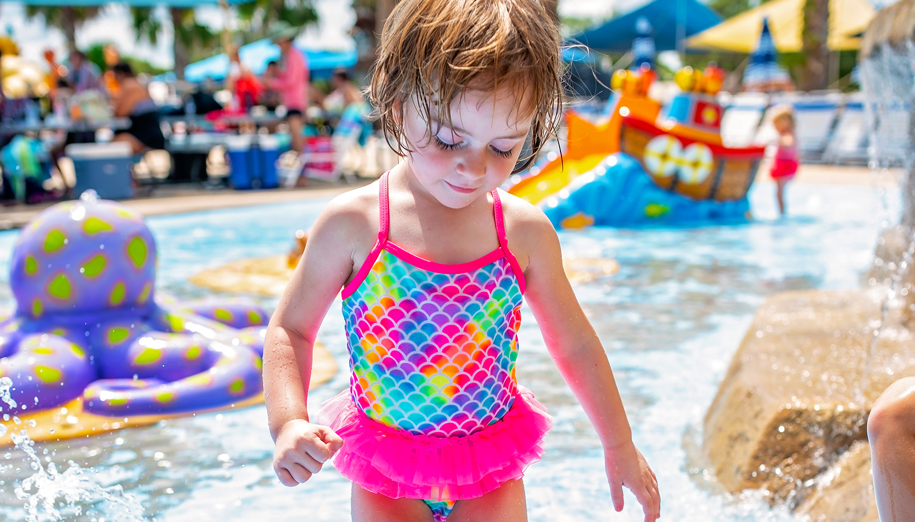 A young child in a colorful swimsuit plays in a shallow water park pool with inflatable toys and other children in the background.