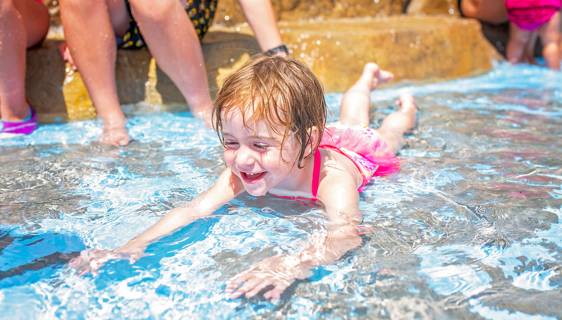 A young child in a pink swimsuit is smiling while lying on their stomach in shallow water at a pool, with other people sitting on the edge in the background.