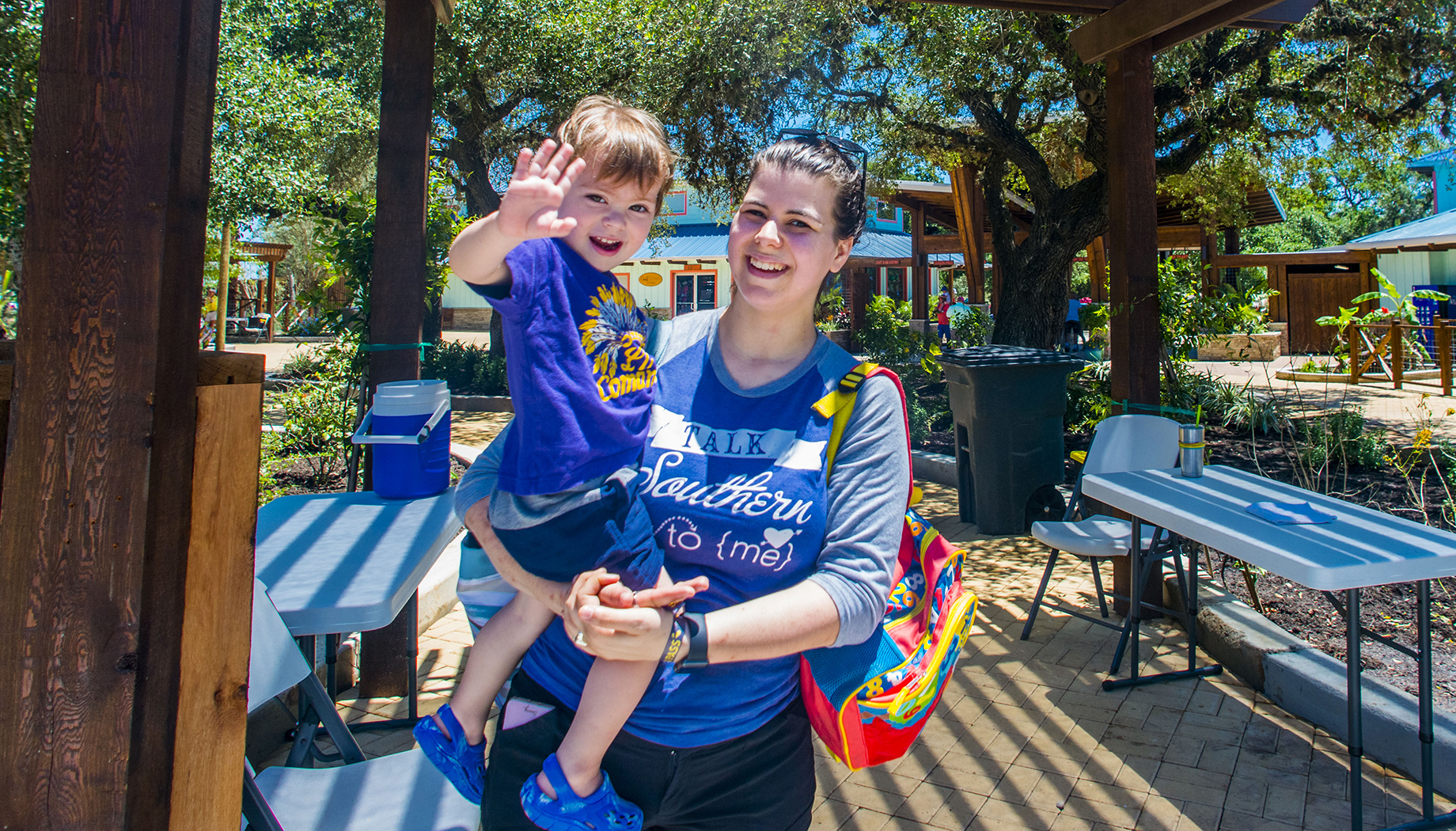 A woman holding a young child who is waving, both standing outdoors on a sunny day with tables and chairs around.
