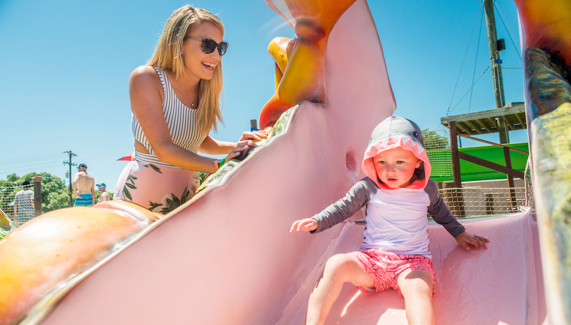 An adult assists a child riding down a colorful slide on a sunny day. The child wears a hooded shirt and shorts, and the adult is wearing sunglasses and a striped top.