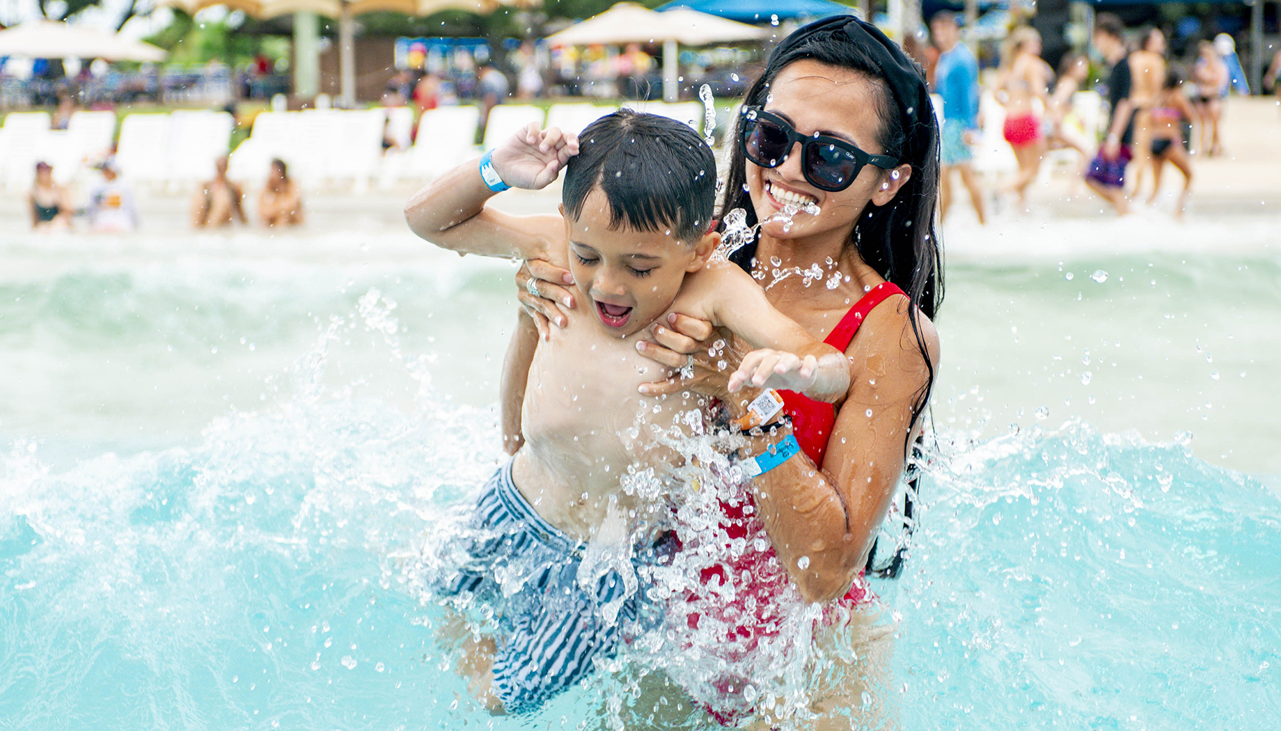 A woman in a red swimsuit lifts a young boy out of the water at a beach. Both are smiling, and there are other people and beach chairs in the background.