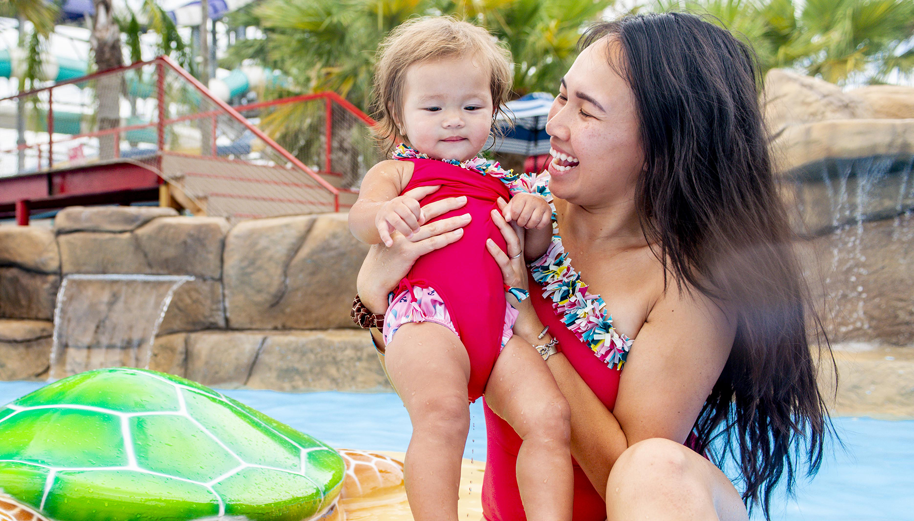 An adult and a toddler in matching red swimsuits are enjoying a splash pad with a green turtle water feature in the background.