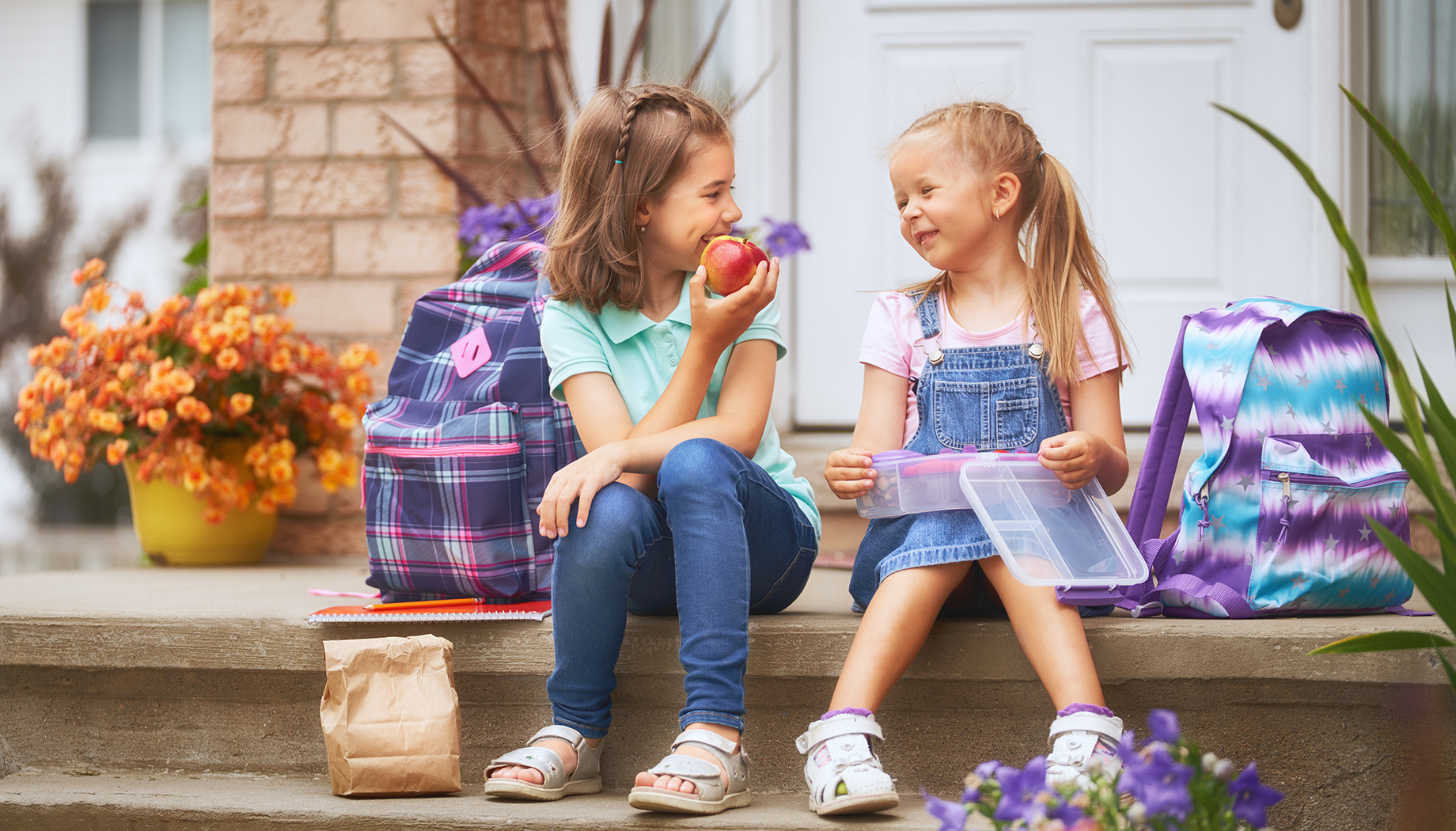 Two girls on a front step with purple and blue backpacks. They are eating an apple and lunch.