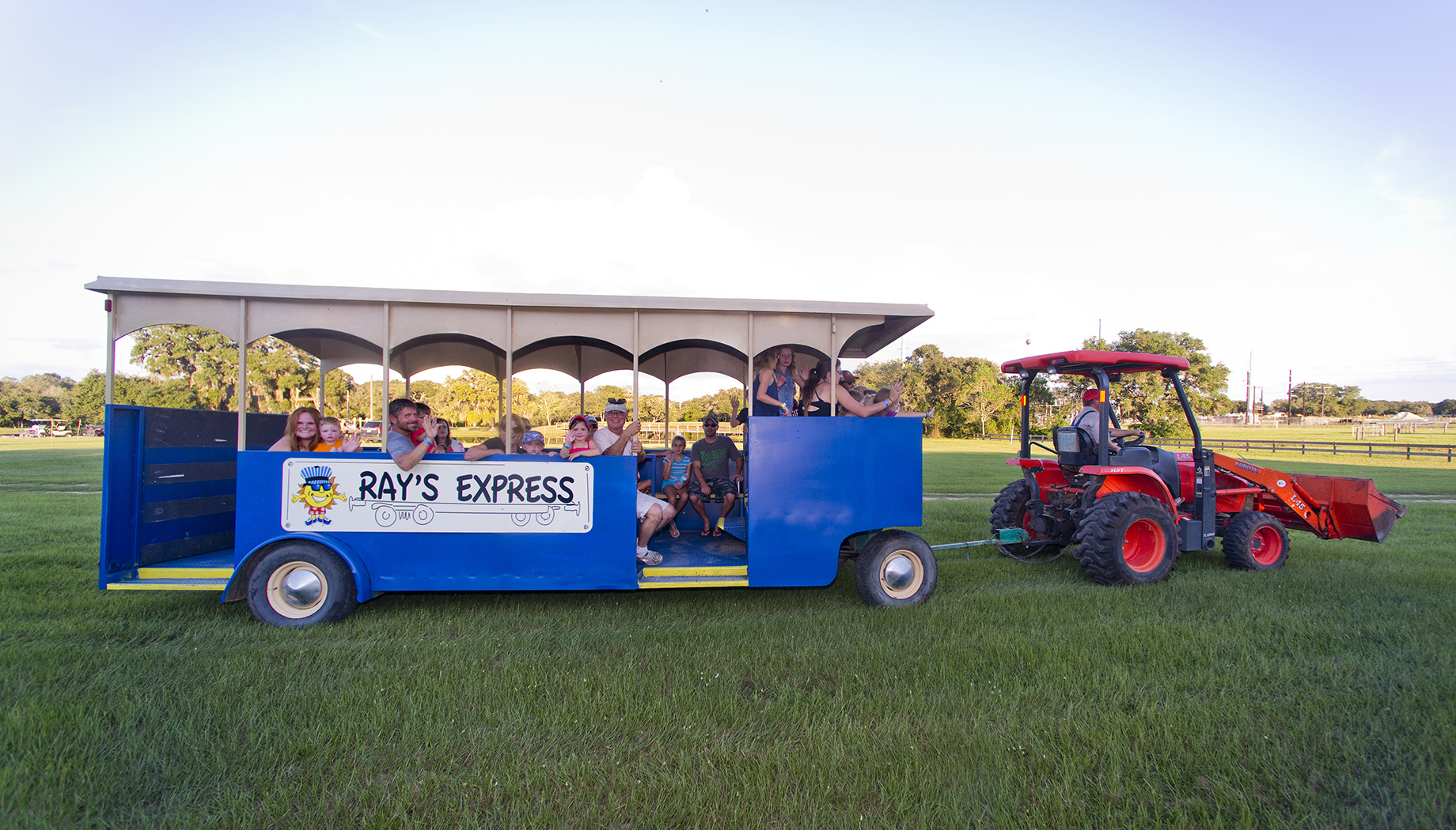 A red tractor pulls a blue open-sided trolley labeled "Ray's Express" across a grassy field with passengers on board.