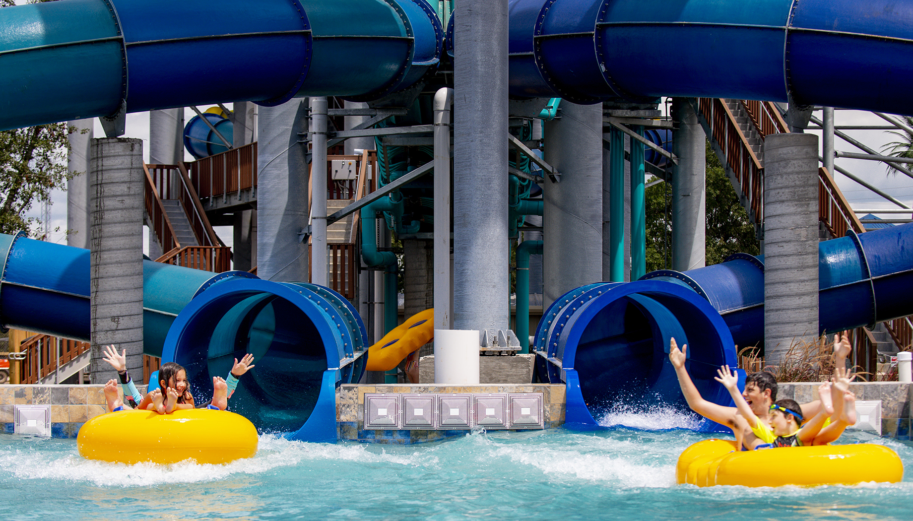 People riding yellow inner tubes emerge joyfully from water slides into a pool at an outdoor water park.