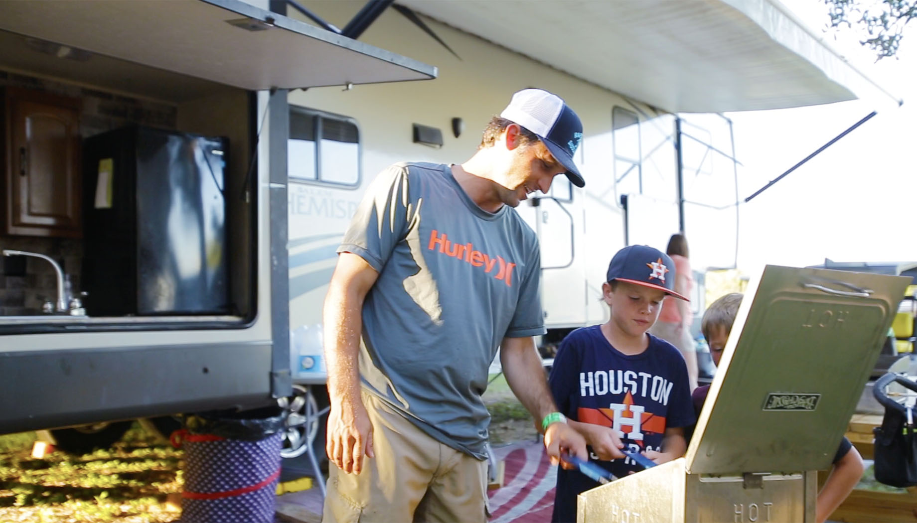 A man and two boys are looking at a grill near an RV. The man is wearing a gray shirt and cap, and the boys are in dark caps and shirts.
