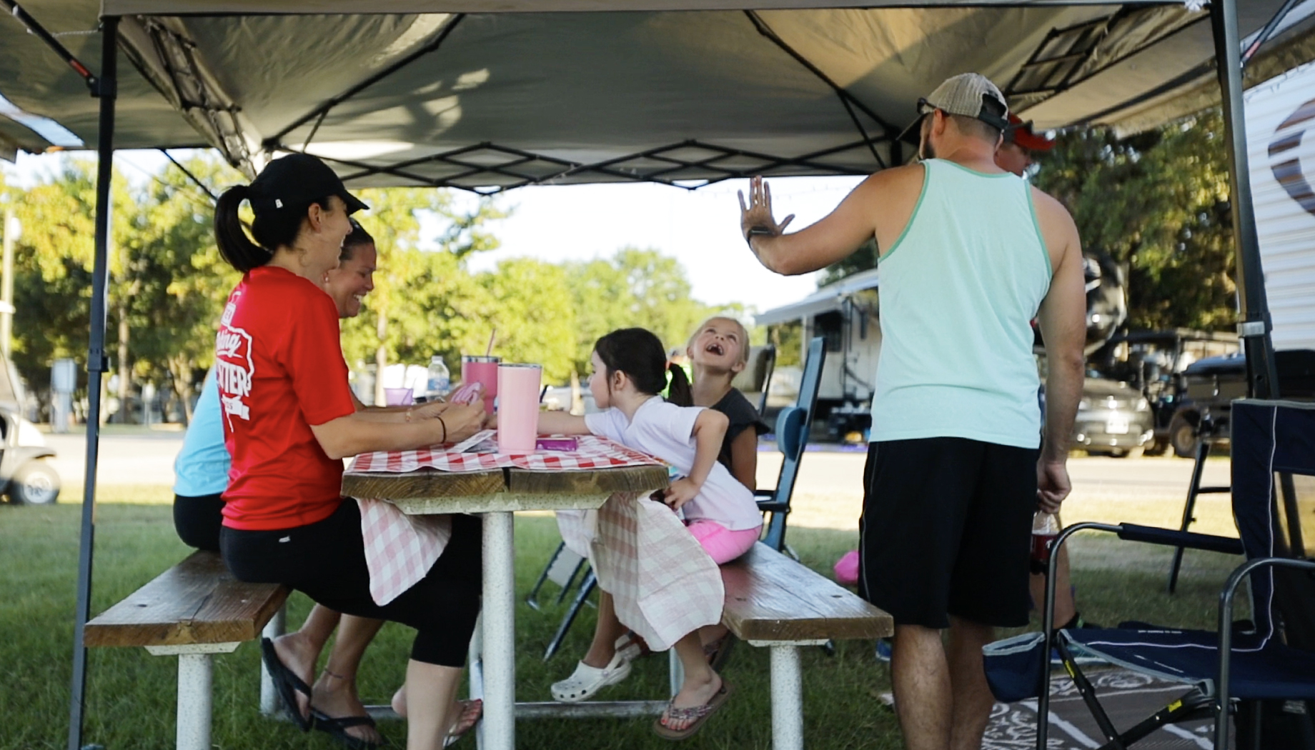 A group of people sit around a picnic table under a canopy, with a man standing and talking, and a child laughing enthusiastically.