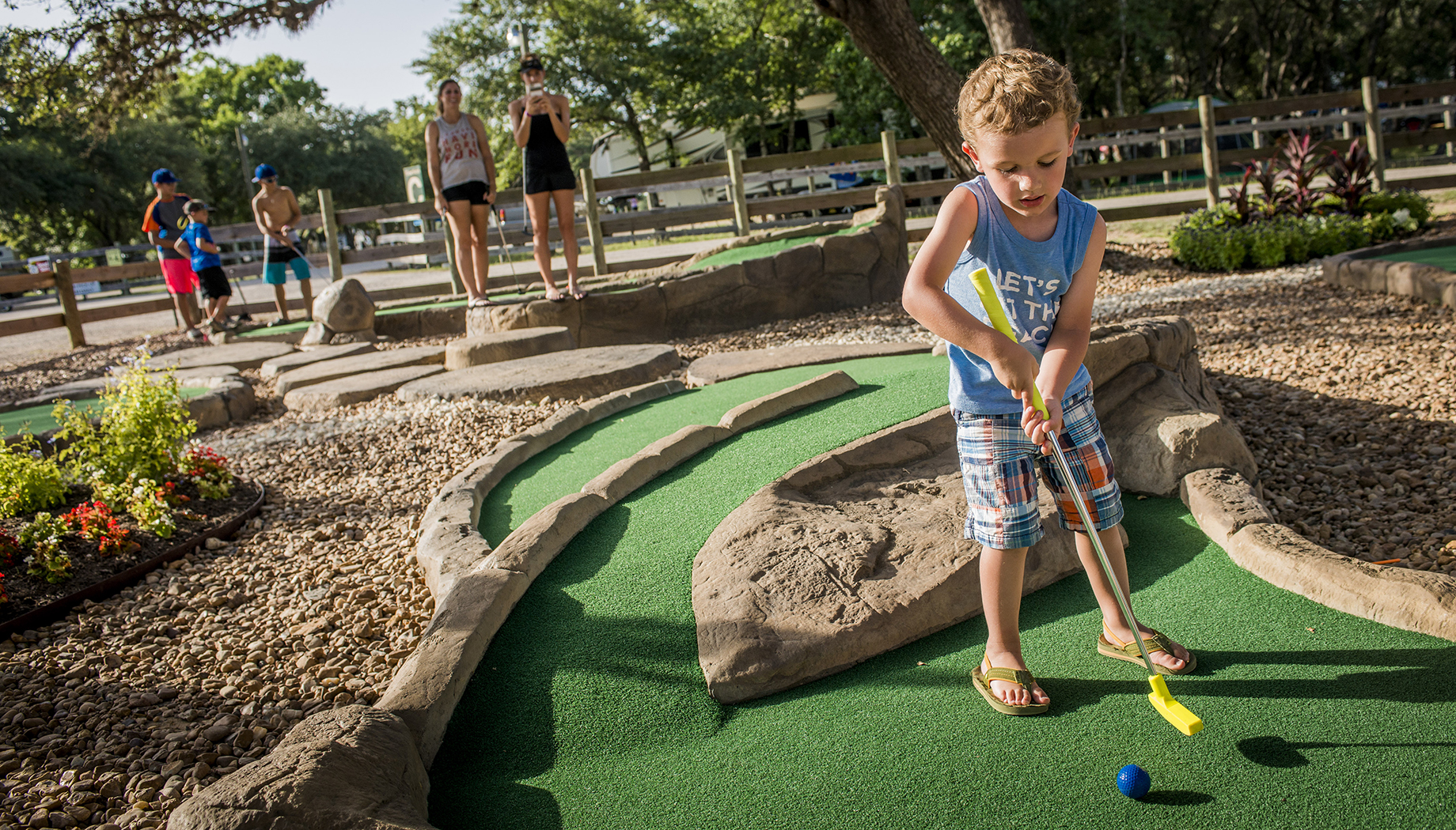 A young boy wearing plaid shorts and a blue tank top plays mini golf on a course with rocks, as other people watch in the background.