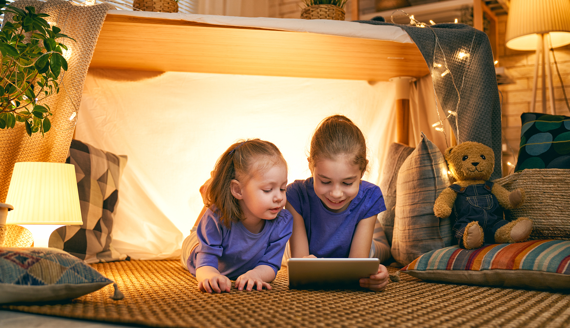 Two children wearing blue shirts are lying on the floor inside a cozy blanket fort, illuminated by string lights, looking at a tablet.