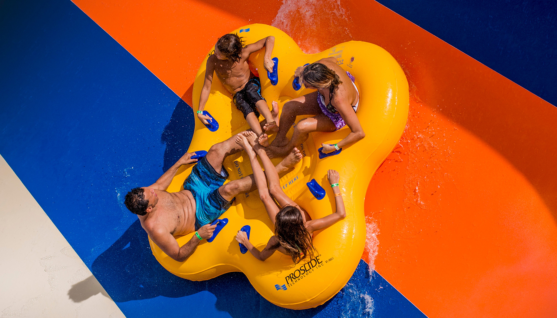 Four people ride an inflatable raft down a water slide, holding onto blue handles. The slide has bright blue and orange sections.