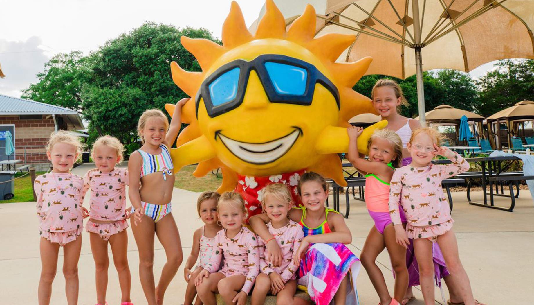 A group of children in swimwear pose with a large sun mascot wearing sunglasses and a floral shirt under a shaded area by a pool.