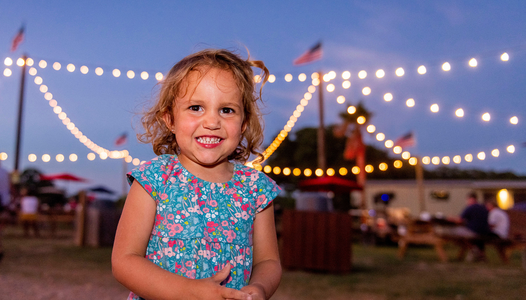 A smiling child wearing a floral dress stands outdoors at dusk with string lights and American flags in the background.