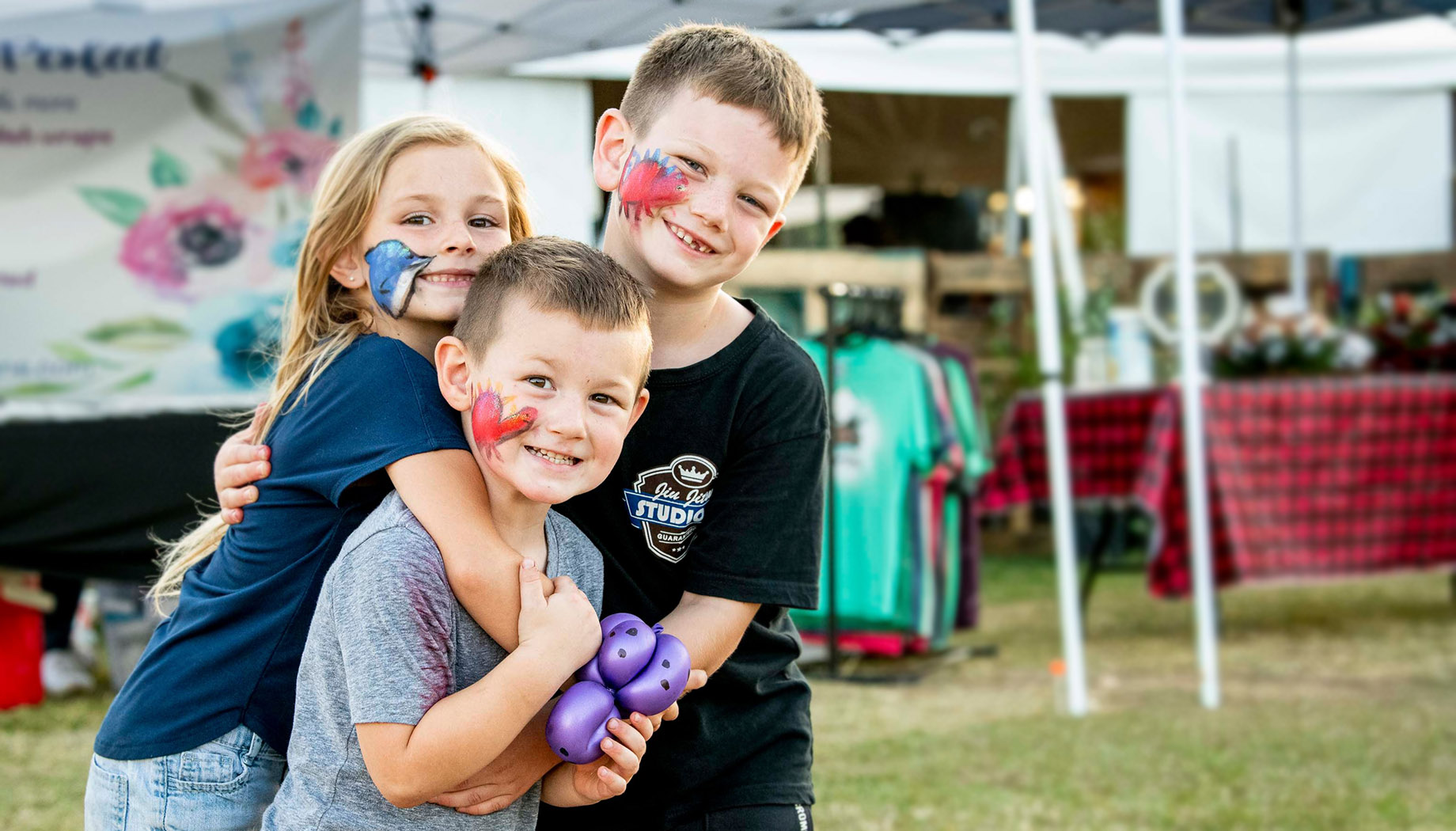 Three children with face paint smile and pose together at an outdoor event. The boy in the front holds a purple balloon animal.