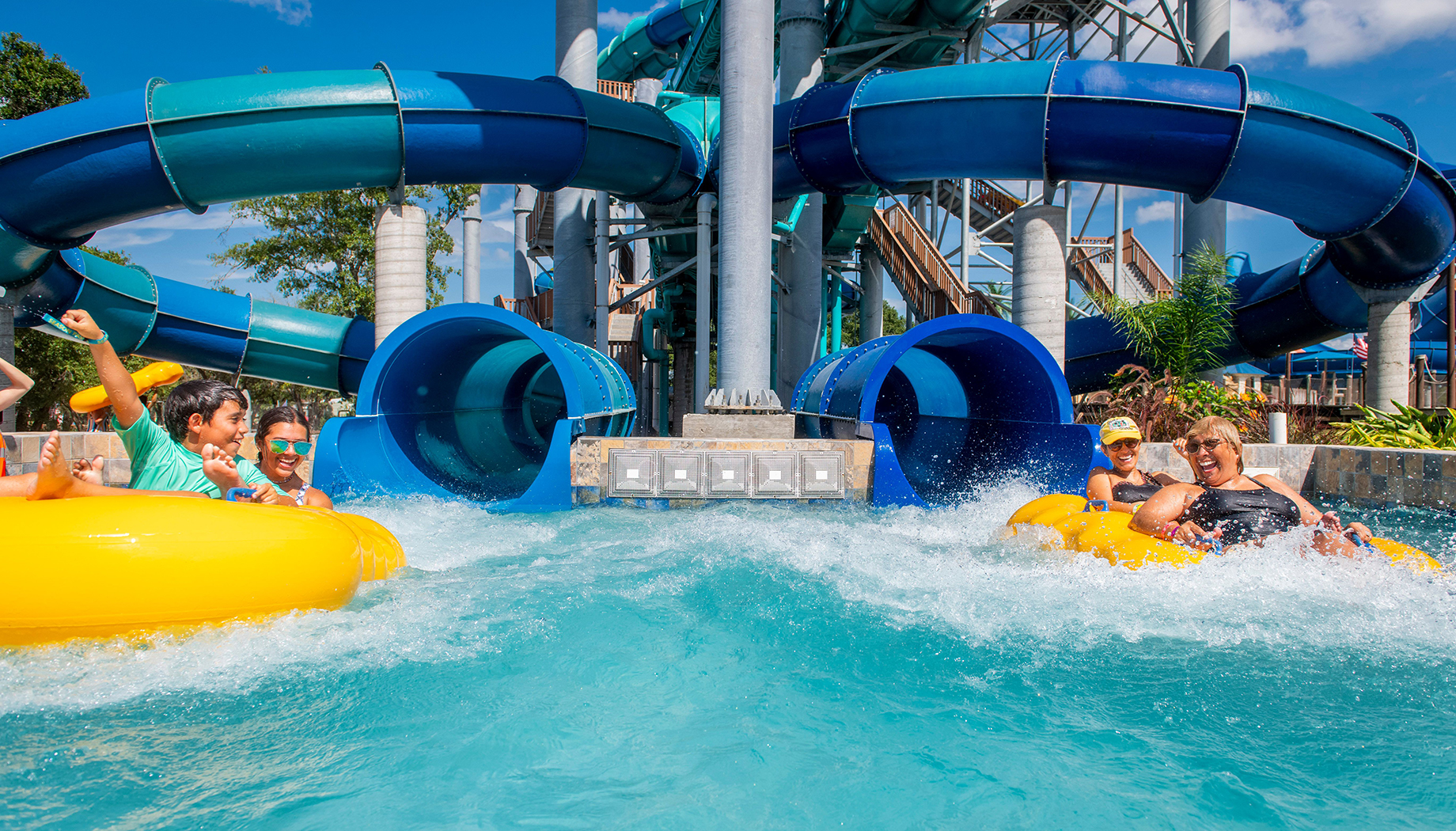 People riding yellow and blue inner tubes down a water slide at a water park with a backdrop of twisting water slide structures and clear blue sky.