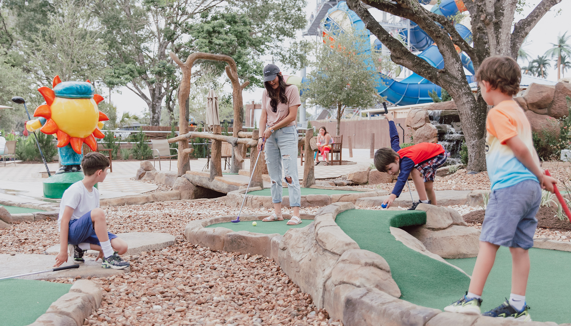 A group of children and an adult playing mini-golf at an outdoor course with a colorful, playful setting.