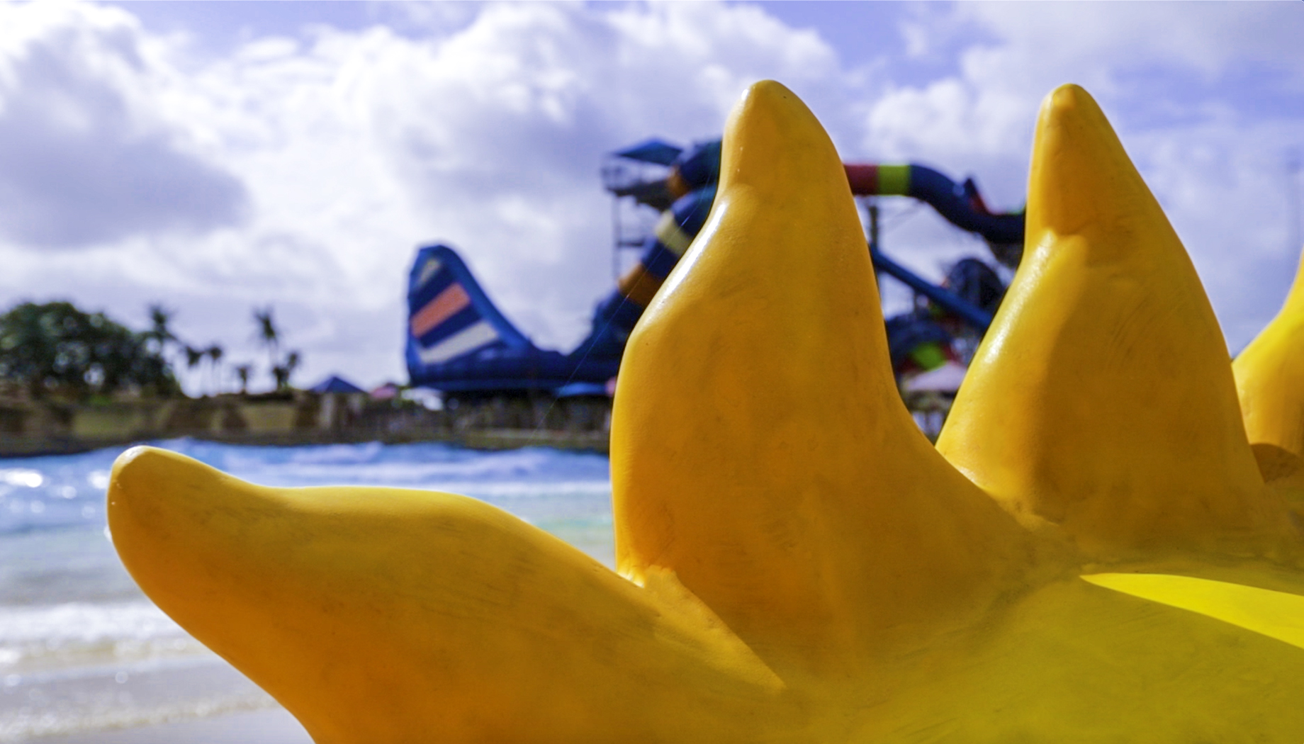 Close-up of a yellow sun-shaped sculpture with a water park featuring colorful slides in the background against a partly cloudy sky.