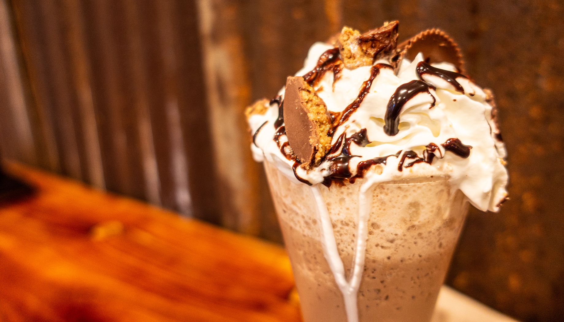 A tall glass of milkshake topped with whipped cream, chocolate drizzle, and pieces of peanut butter cups sits on a wooden table.