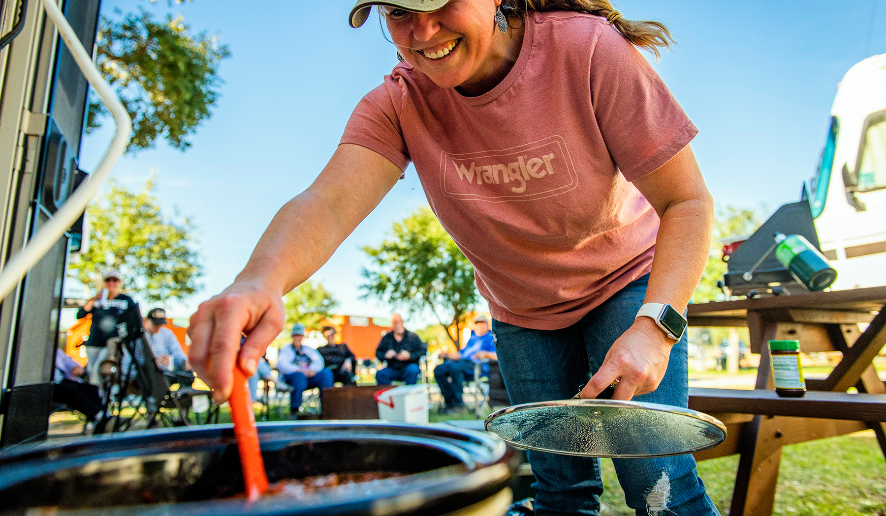 Woman stirring chili in a crockpot with an RV and people in the background.