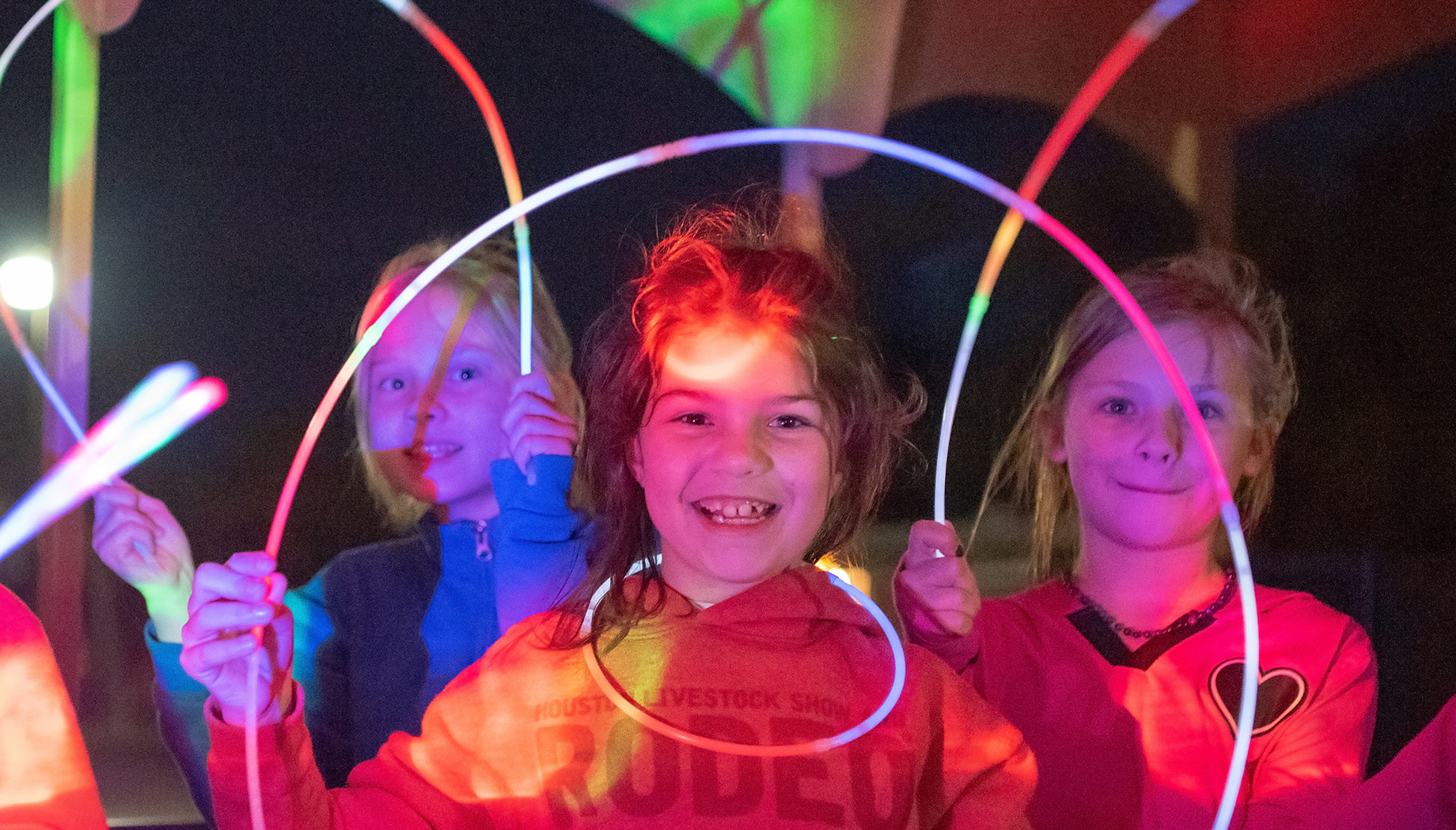 Three children smiling and holding colorful neon light sticks in a dark setting.