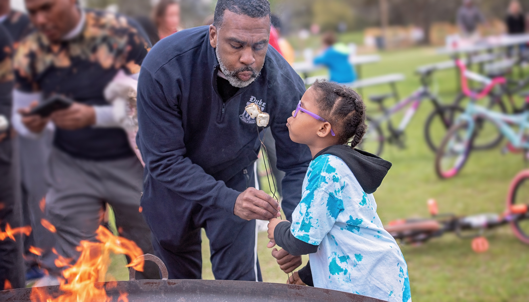 A man helps a child roast marshmallows over an outdoor fire pit at a park during the day, with bicycles and picnic tables in the background.
