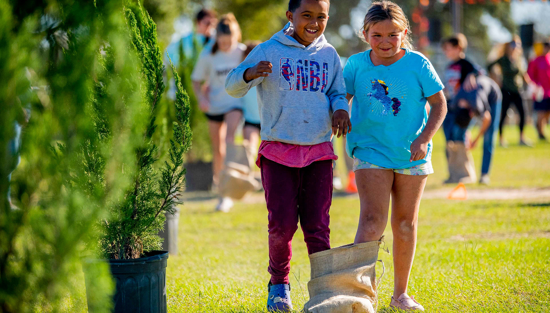Two children participate in a sack race during an outdoor event, both smiling and holding hands, surrounded by greenery and other participants in the background.