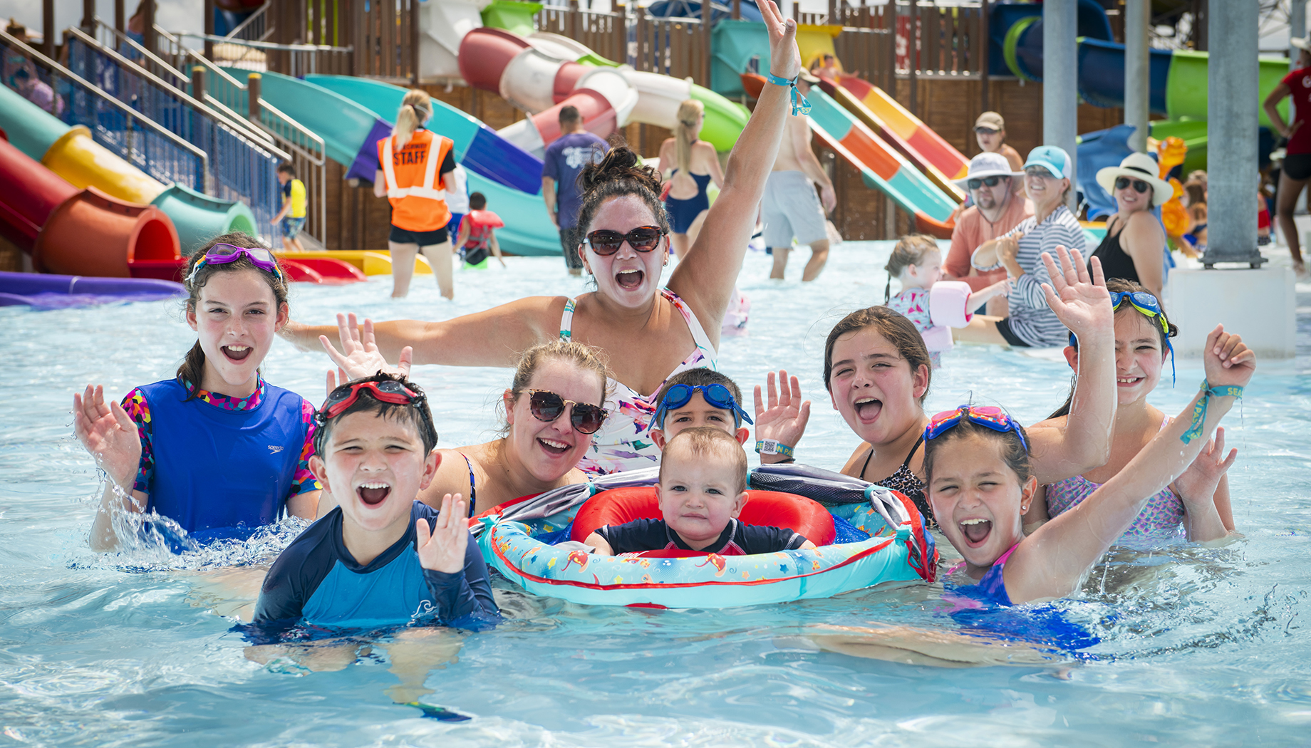 A group of people, including children and adults, are enjoying themselves in a water park pool. They are all smiling and waving, with colorful water slides visible in the background.