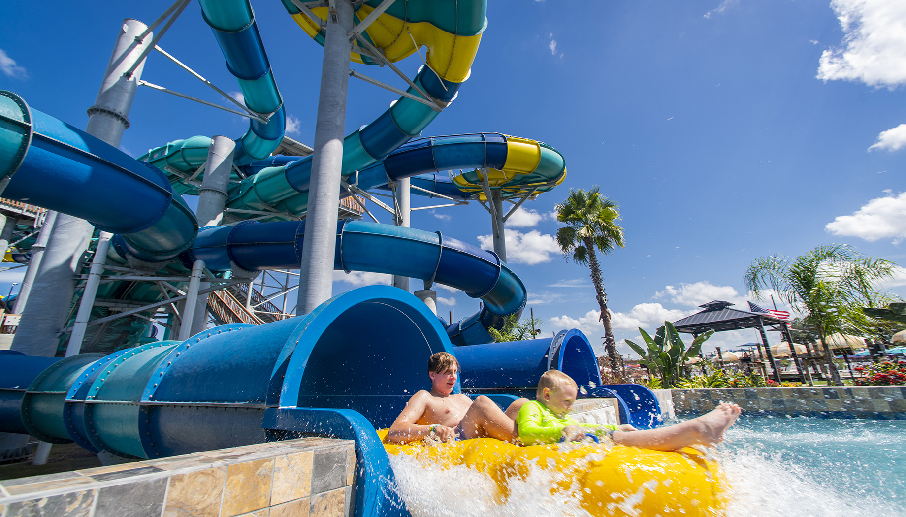 Two children on a yellow inflatable raft exit a blue water slide into a splash pool at a water park under a clear blue sky.