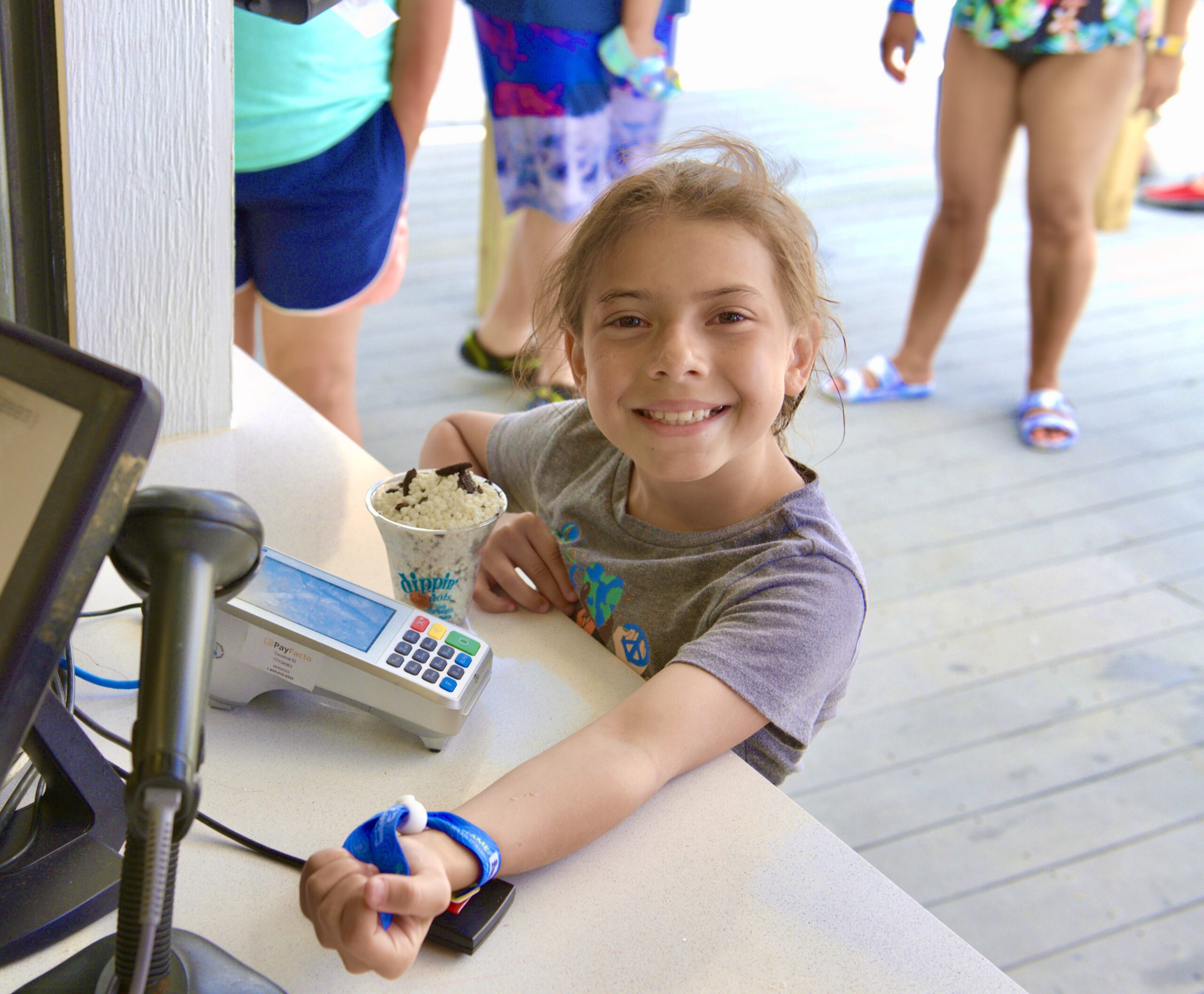Young girl smiling and holding her wristband up while standing at a counter with a card reader. She has a dessert bowl filled with ice cream and toppings next to her.