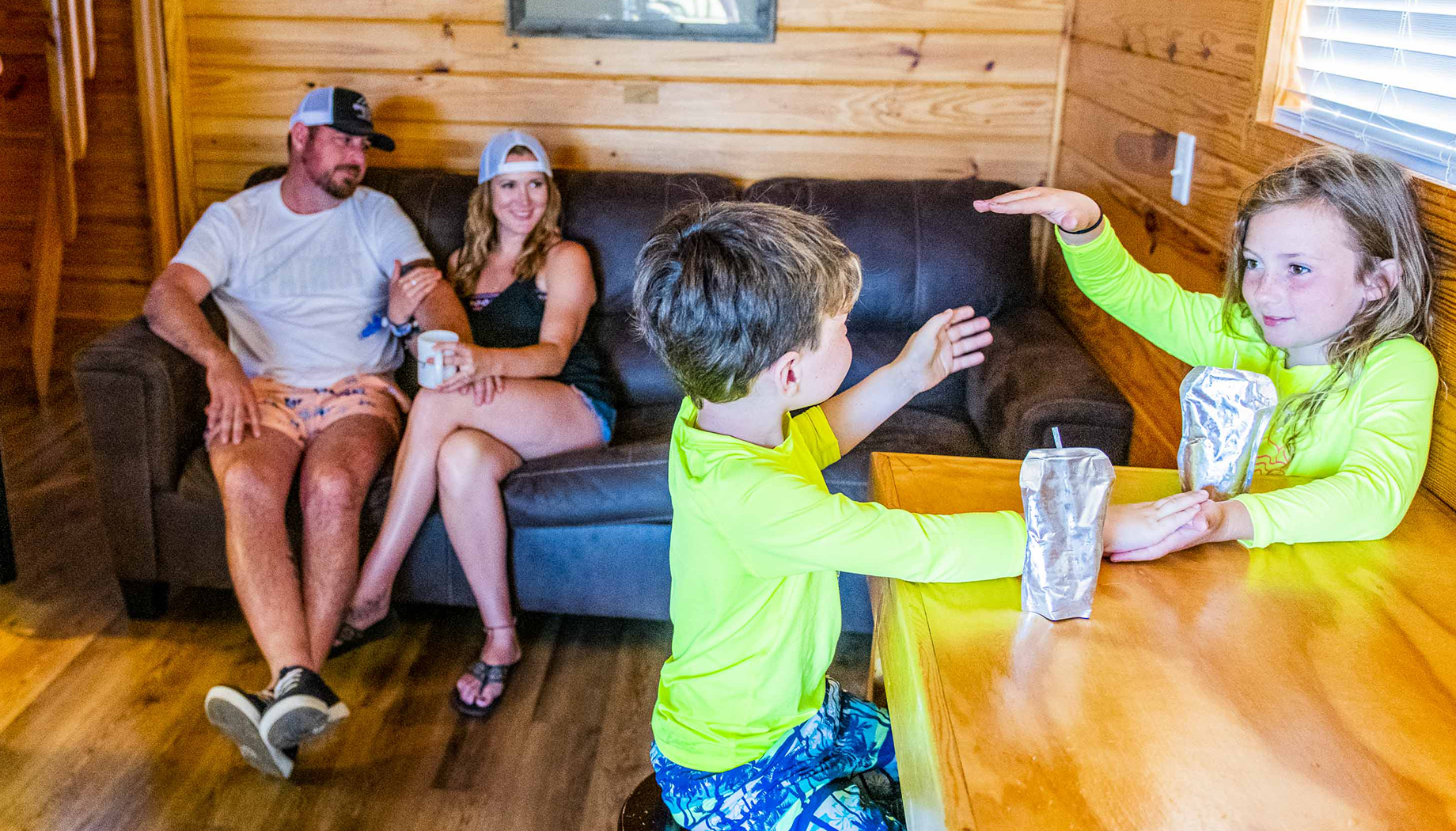 Two children in swimwear sit at a wooden table, playing and drinking from pouches. Behind them, two adults relax on a couch in a wood-paneled room.