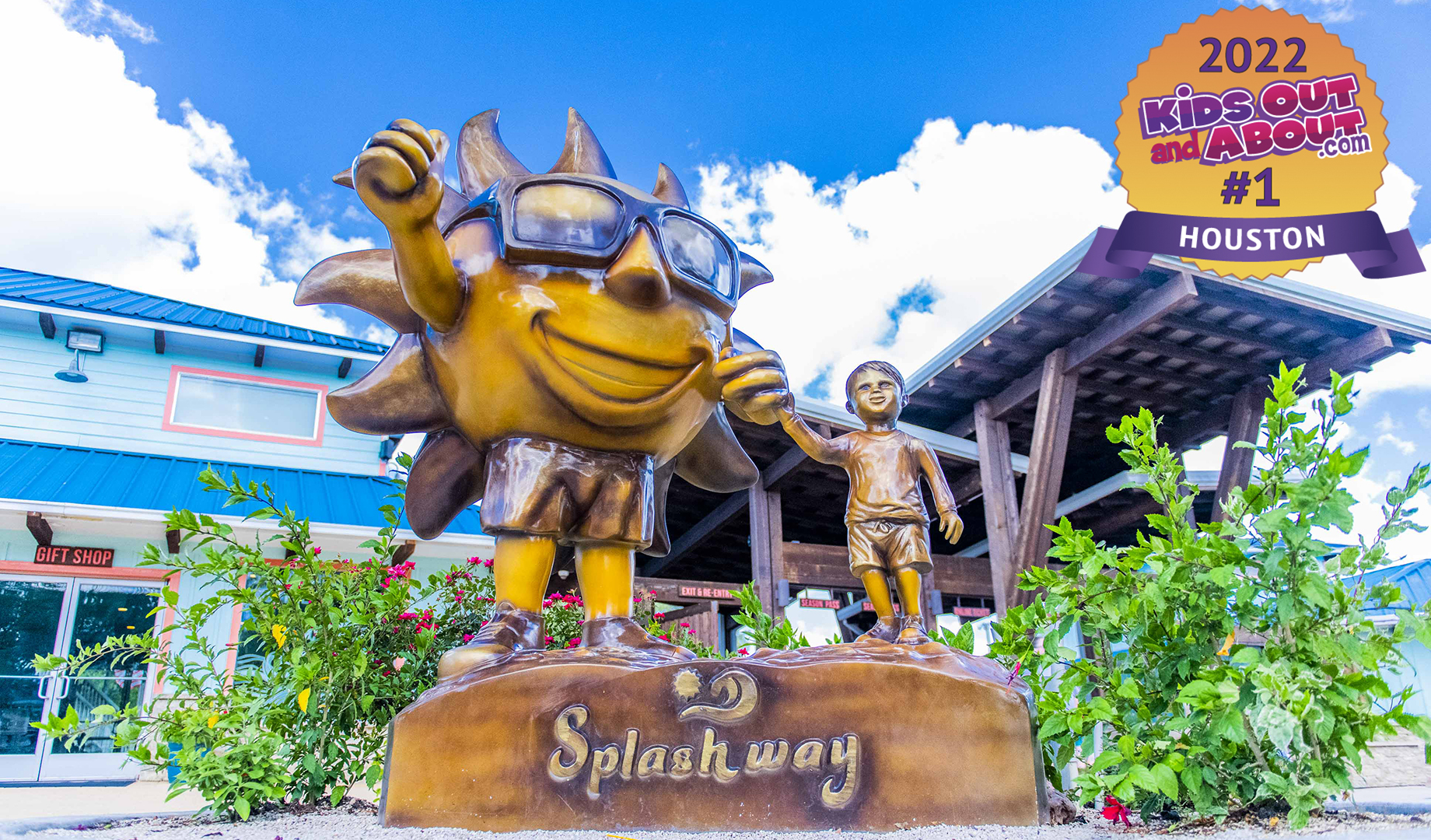 Bronze statue of a sun character and a child holding hands, situated outdoors near a building with a blue roof. A sign displays "2022 Kids Out and About #1 Houston.
