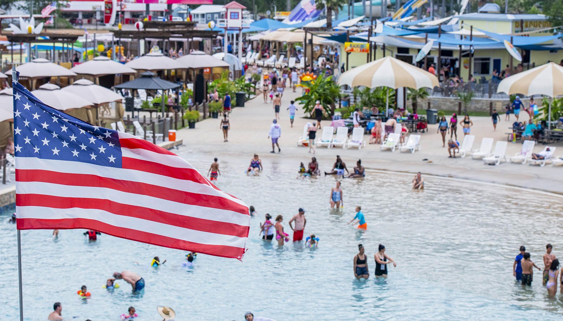 A large American flag flies over a crowded water park, with people swimming, sunbathing, and walking around. Various water rides and concession stands are visible in the background.