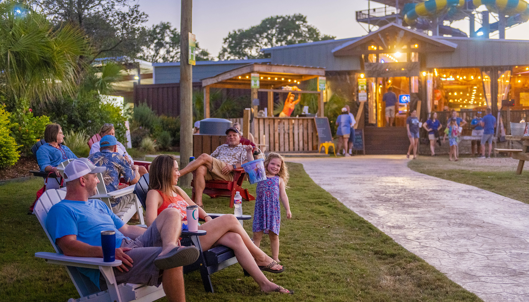 A group of people relaxes on lawn chairs outside a well-lit building at dusk. A young girl stands smiling in the center, and people can be seen conversing and moving in the background.
