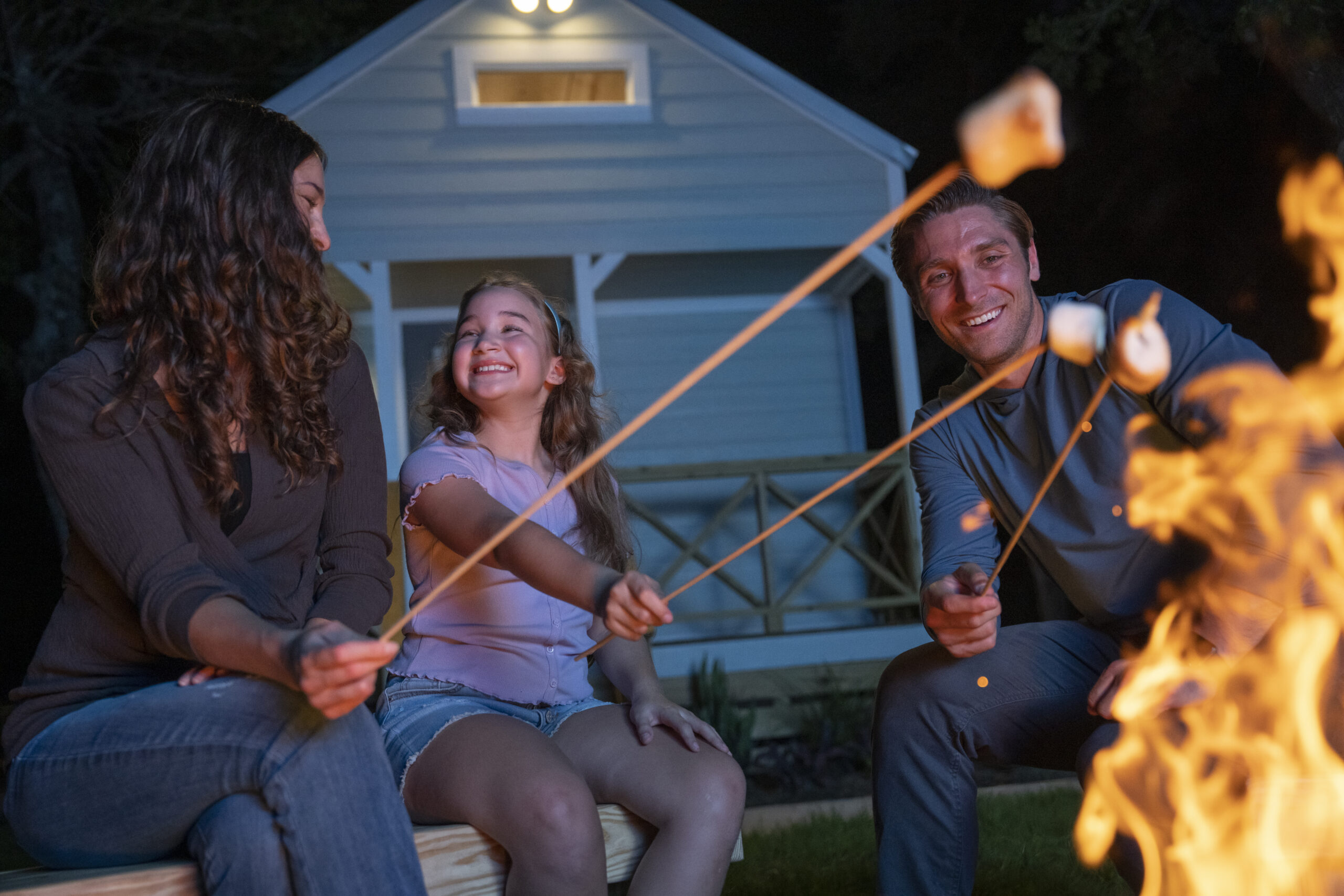 Family toasting marshmallows over a campfire with a cabin in the background.