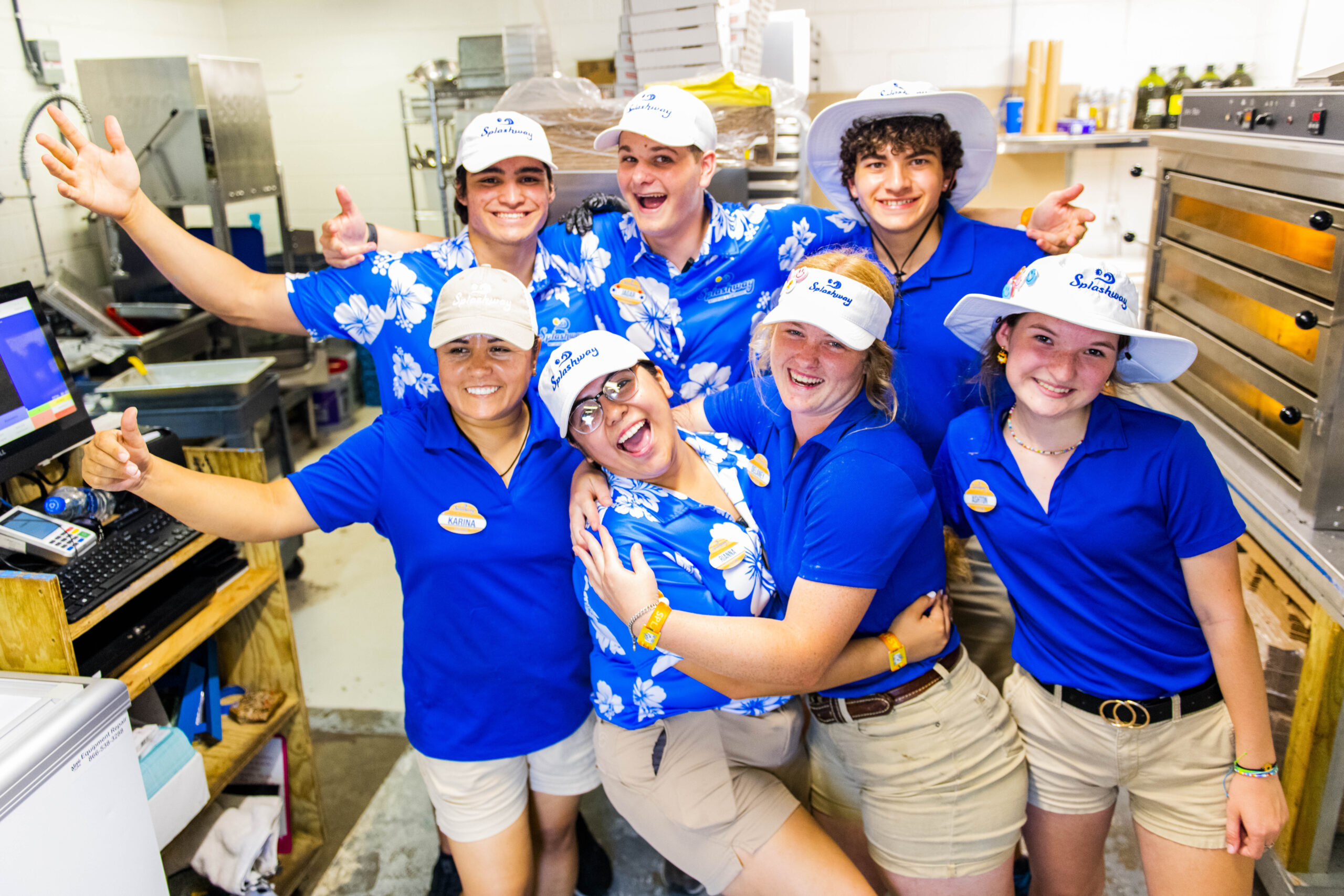 A group of seven people wearing blue shirts, beige shorts, and white hats with a restaurant logo stand together in a kitchen, smiling and making various cheerful gestures.