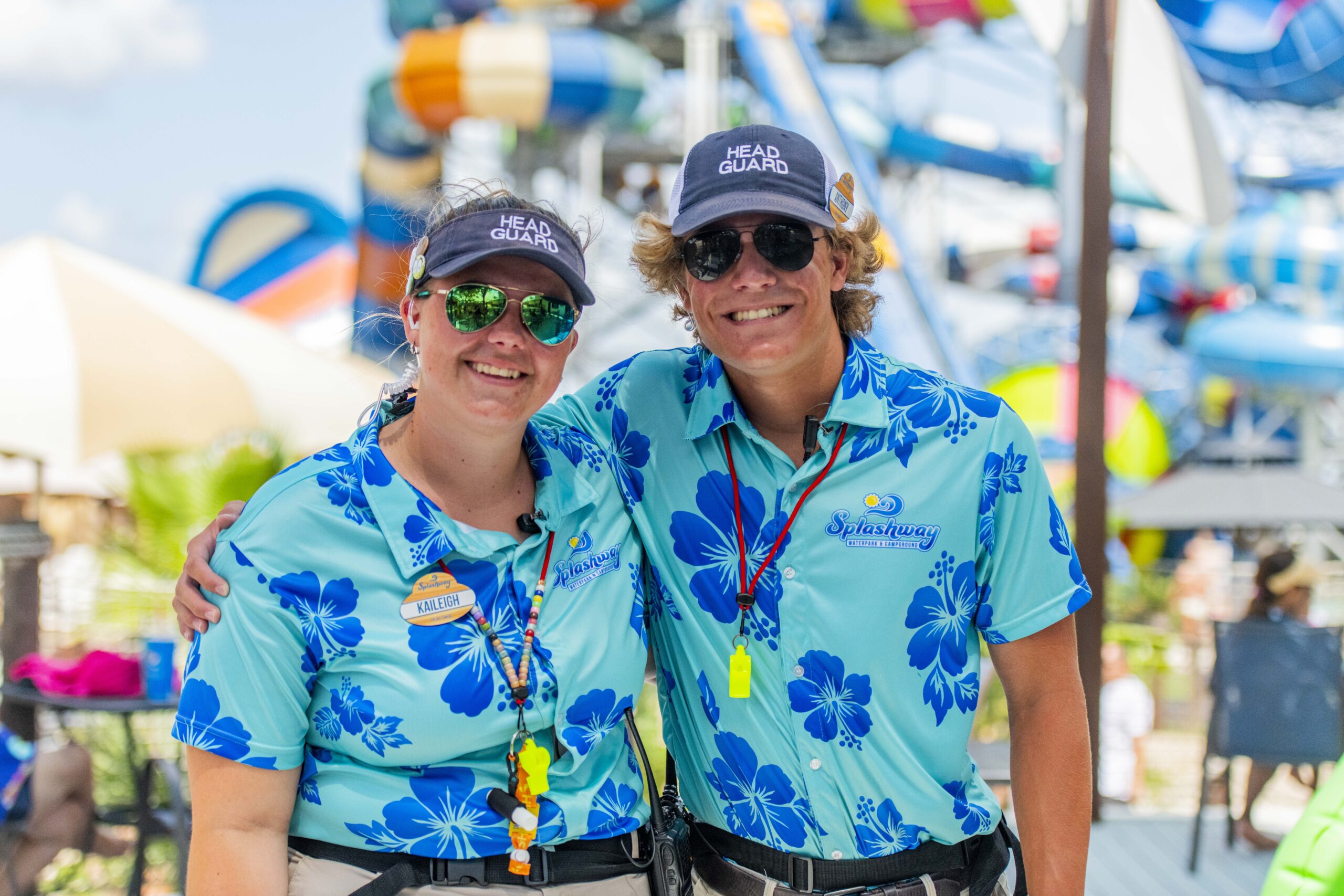 Two lifeguards wearing blue floral shirts and head guard caps smile while standing in front of water park attractions.