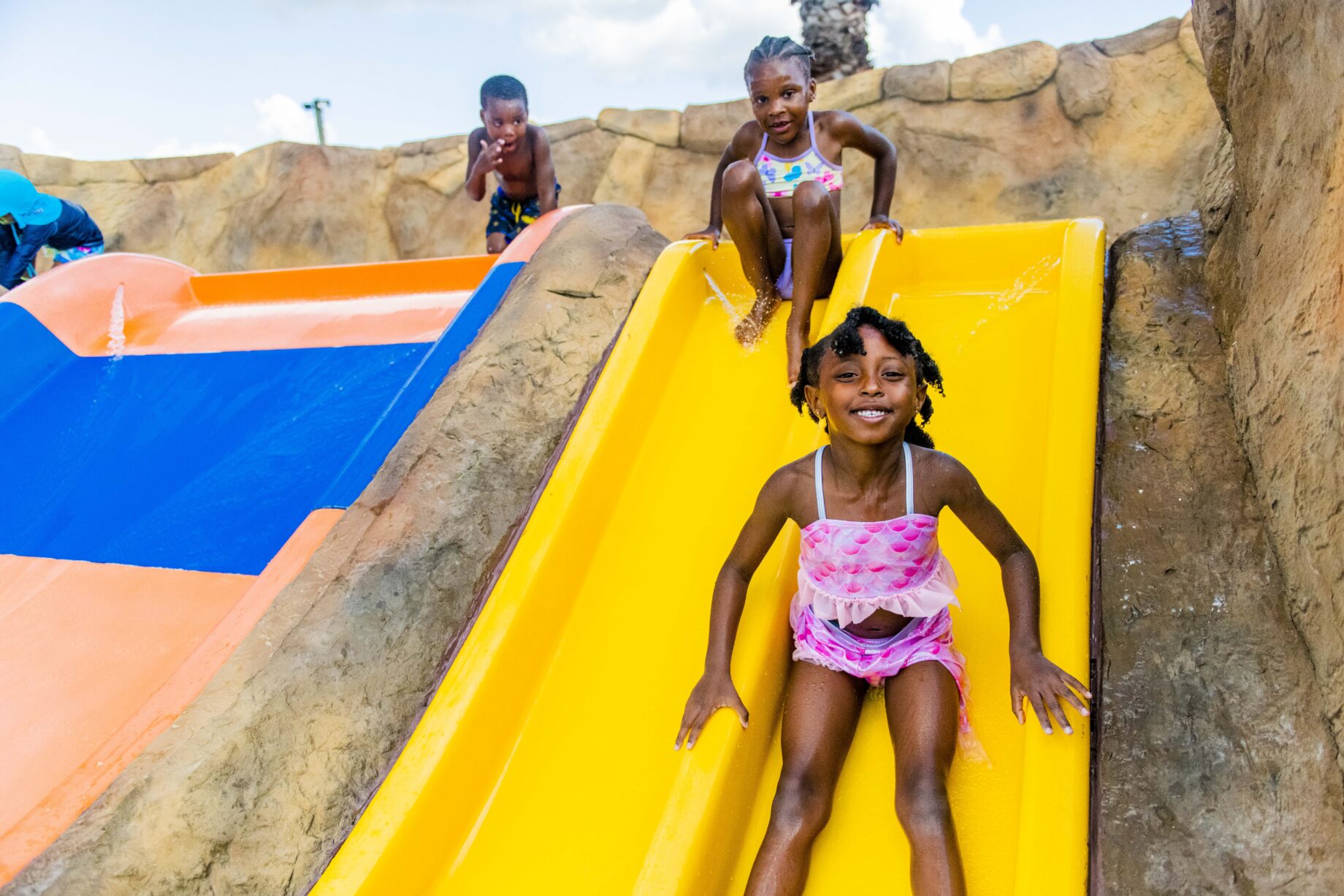 Children in swimsuits slide down yellow and orange slides at a water park. They are smiling and enjoying themselves on a sunny day.