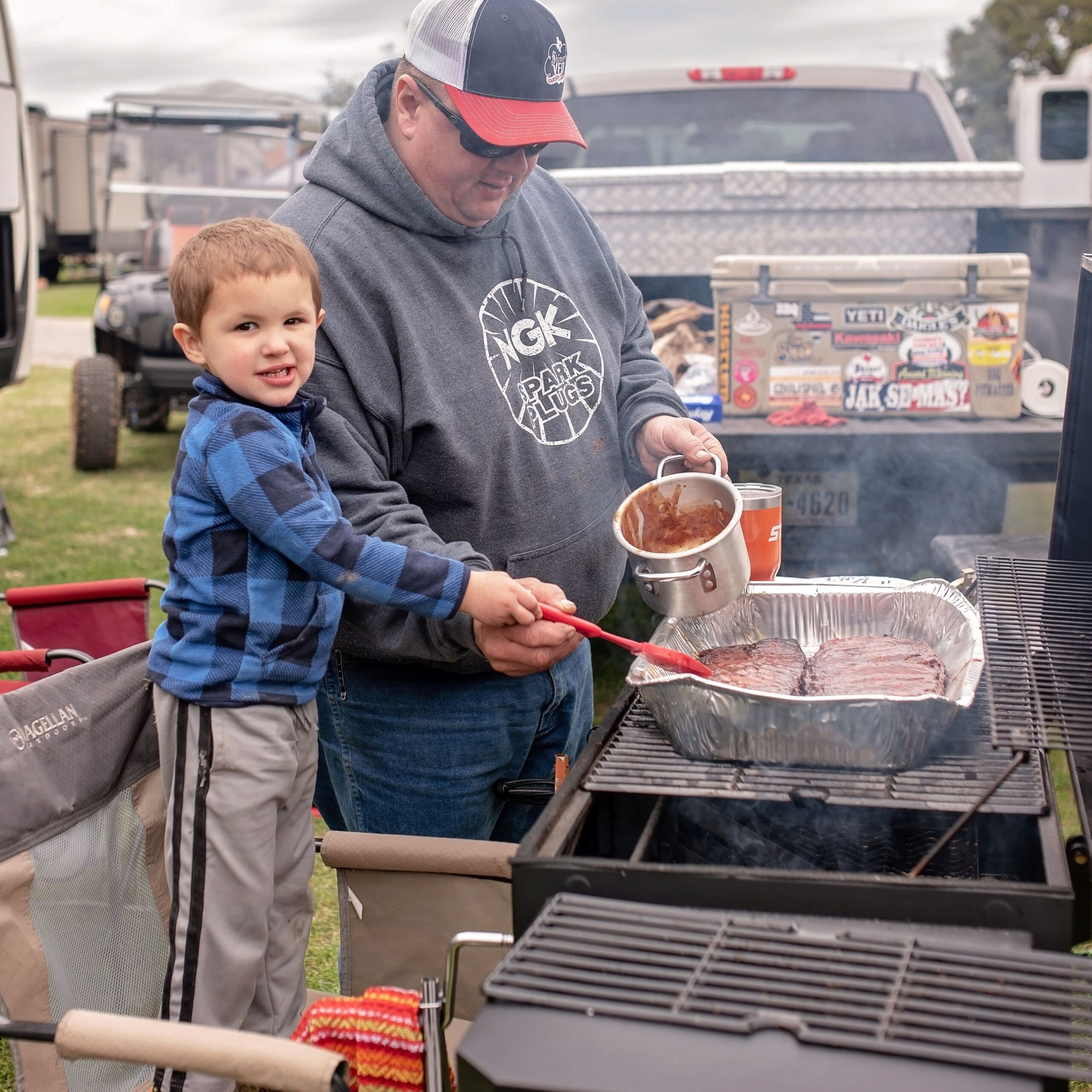 A man helps a young boy pour sauce over meat on a grilling barbecue during an outdoor event. The grill is set up next to a truck with various stickers.
