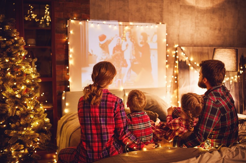 A family of four, dressed in matching plaid pajamas, watches a holiday movie projected on a screen in a cozy, warmly lit living room decorated with Christmas lights and a Christmas tree.