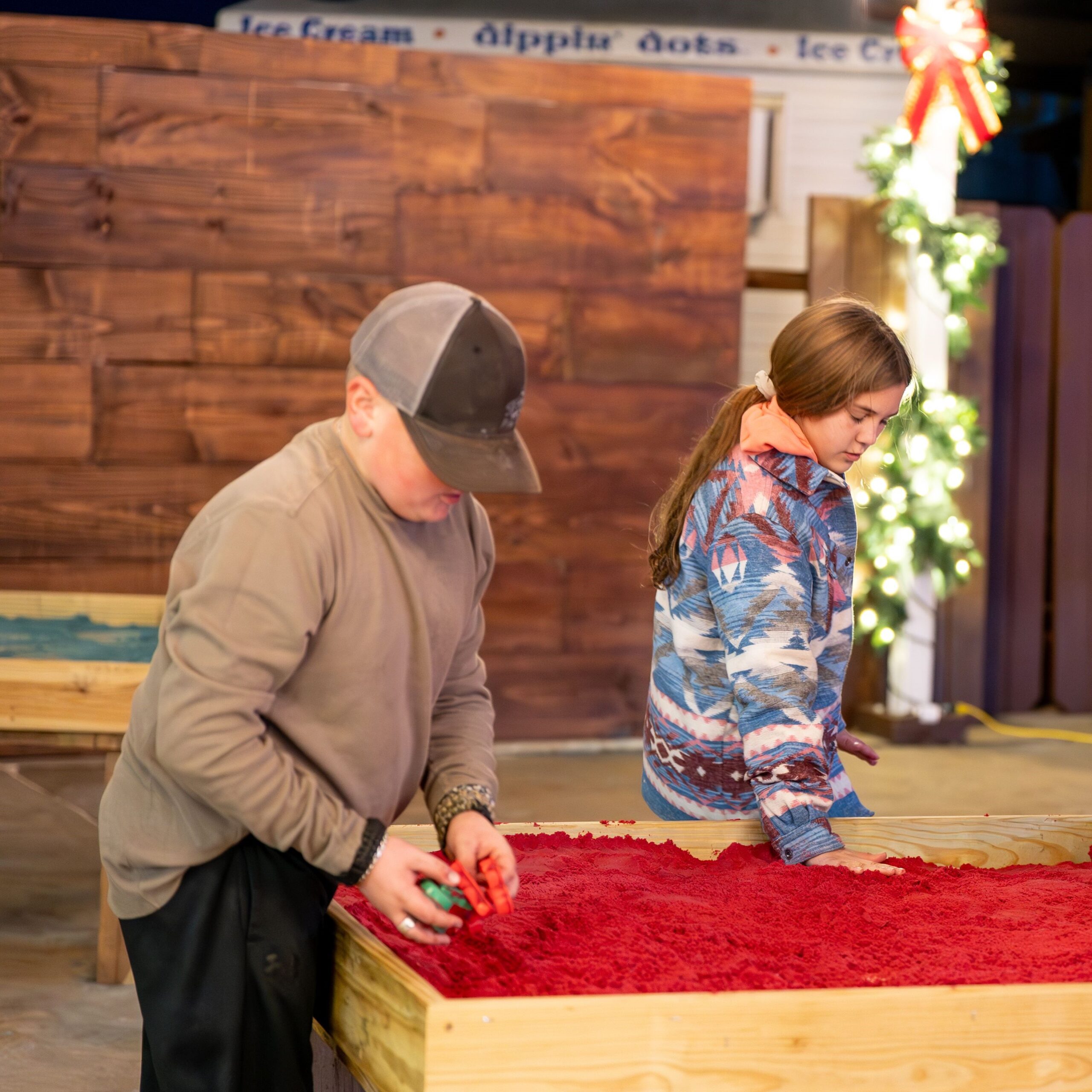 Two children work on a wood project; one boy sands the red surface while the girl stands beside him. A decorated Christmas tree and a wooden wall are visible in the background.