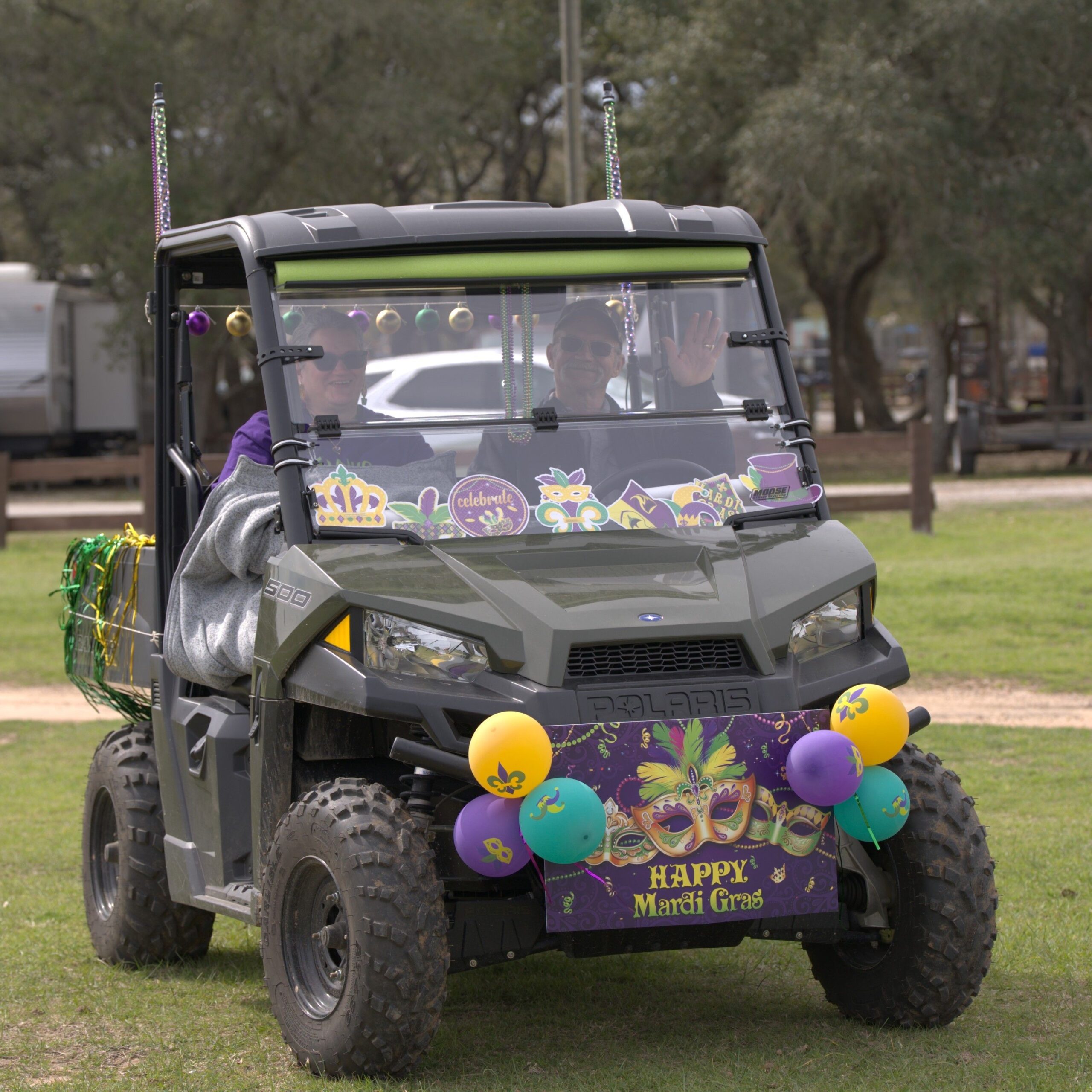 A person drives a decorated off-road vehicle with Mardi Gras banners and balloons on a grass field, waving and wearing sunglasses.